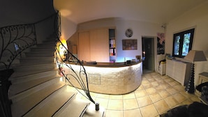 A curved reception desk in a well-lit interior space with a tiled floor, a staircase with ornate metal railings, and a wall-mounted clock. - Auberge Les Arômes (Grasse)