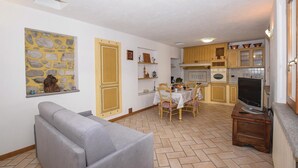 Un espace de vie compact comprenant un canapé, une table à manger avec des chaises, un coin cuisine avec des placards et des appareils électroménagers, et un mur en pierre agrémenté d’un élément décoratif.