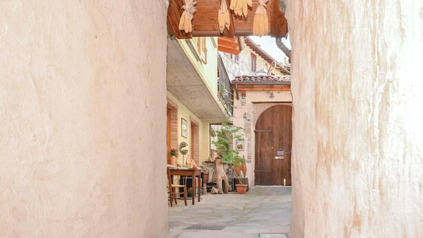 Une ruelle étroite, bordée de bâtiments des deux côtés, avec une porte en bois au fond, et une petite table entourée de chaises devant l’un des bâtiments.