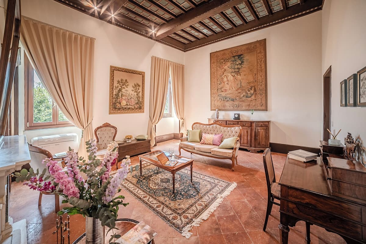 A spacious living room with a wooden coffee table, a sofa, and a dining table. There are curtains on the windows and a large painting on the wall.
