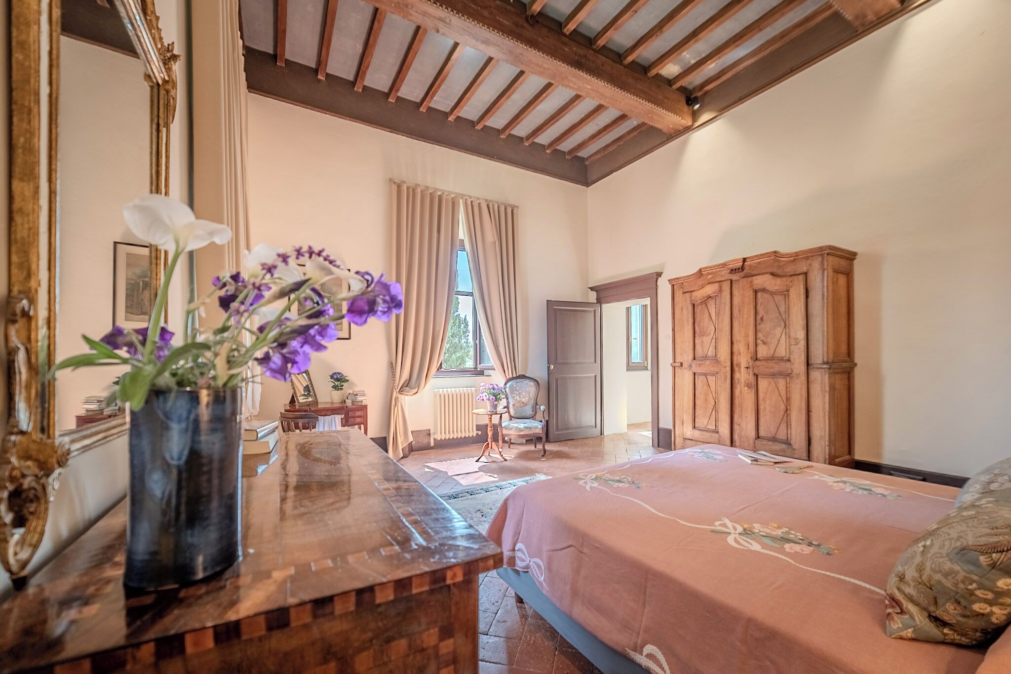 A bedroom with a wooden bed, a wooden dresser, a mirror, and a vase with flowers.
