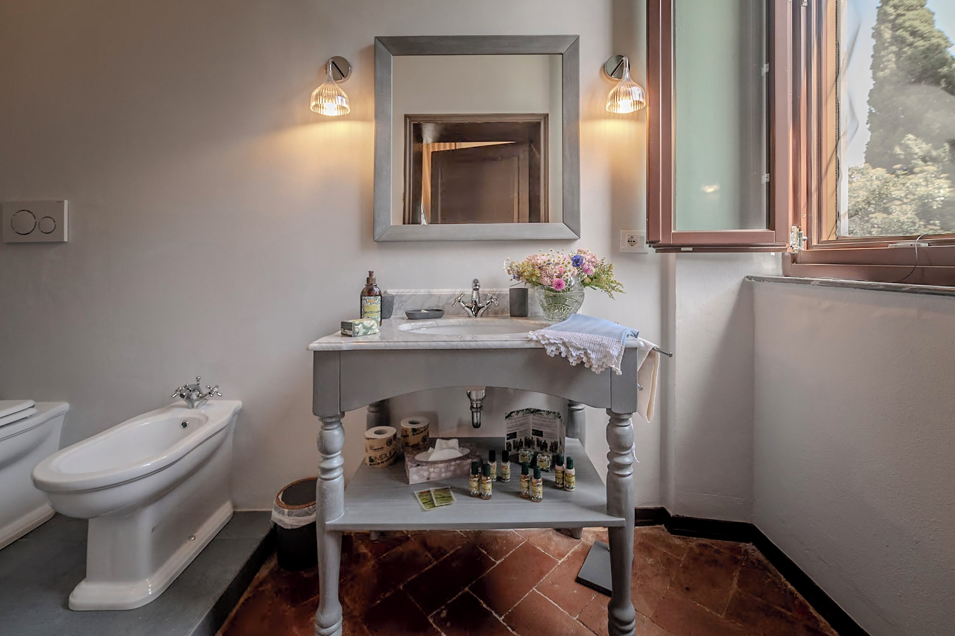A bathroom with a white sink, a mirror, two wall-mounted lights, a window, and a toilet.