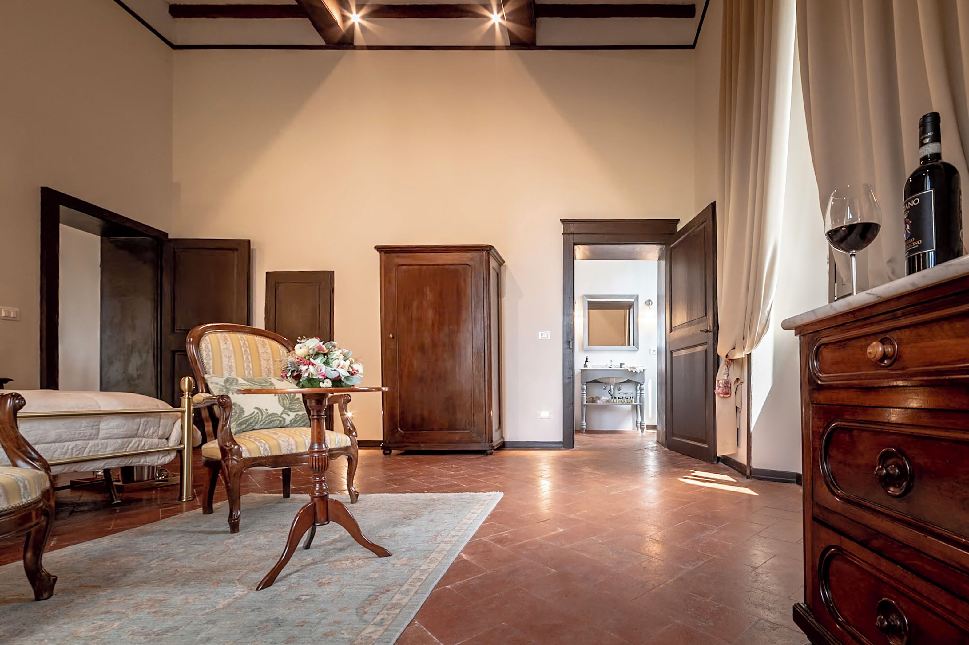 Una habitación amplia con una cama, un armario de madera, una silla con un arreglo floral y un aparador con una botella de vino.