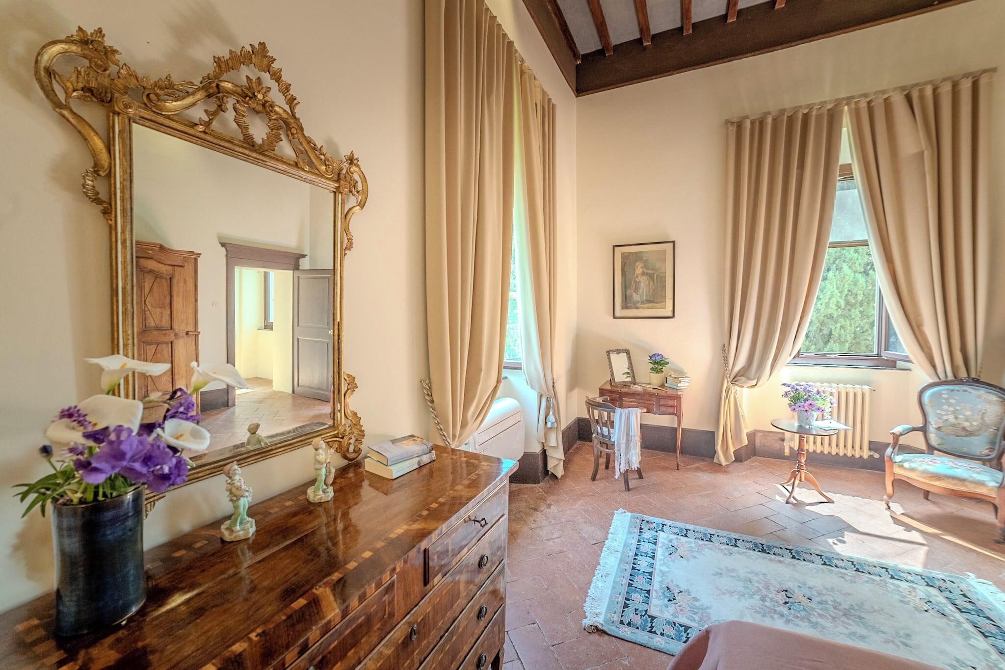 A room with a large ornate mirror, wooden dresser, and a small table with a vase of flowers.