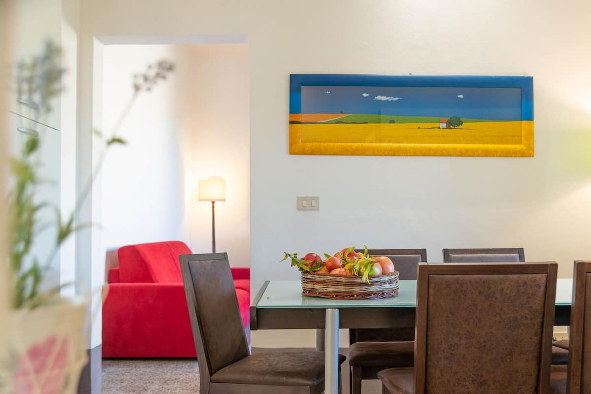 A dining area with a table set for four, a red sofa, and a wall-mounted painting of a landscape.