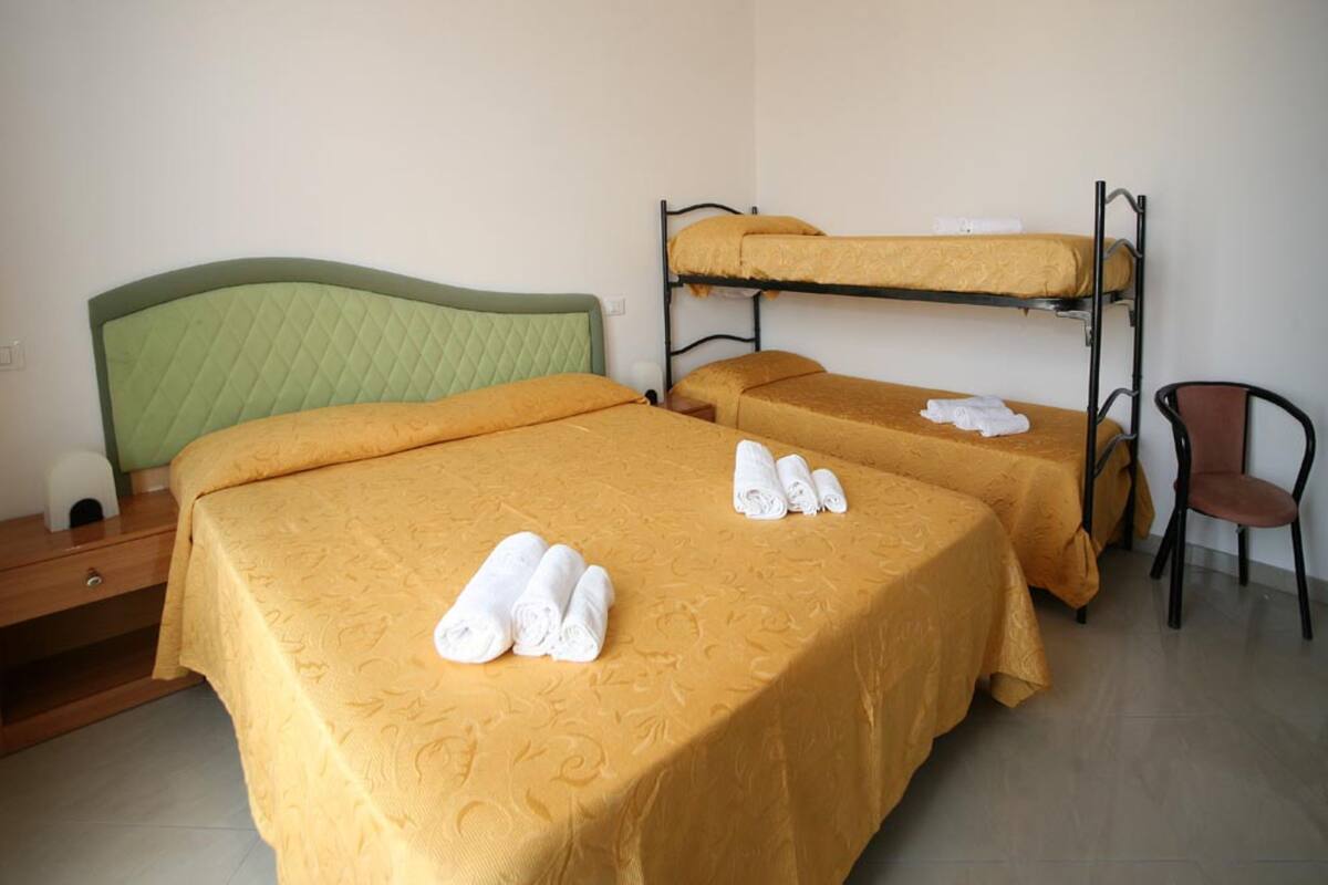 A room with two beds, each with a yellow bedspread and pillows, and a bunk bed above. There are towels on the beds and a chair in the corner.