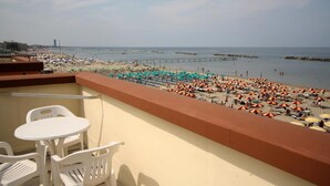 A balcony with white plastic chairs and a table, overlooking a crowded beach with numerous umbrellas and people. - Hotel Riposo (Gatteo)