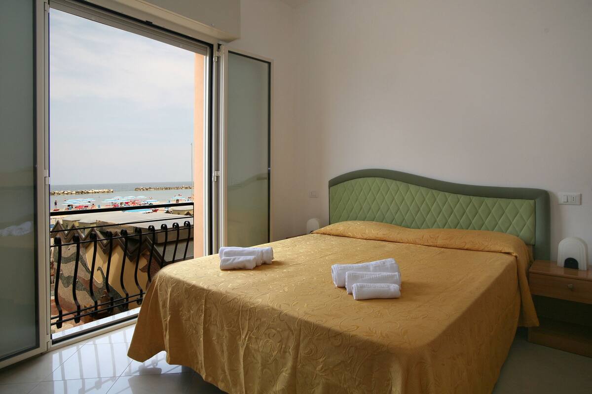 A hotel room with a bed, a nightstand, and a view of the beach through a sliding door.