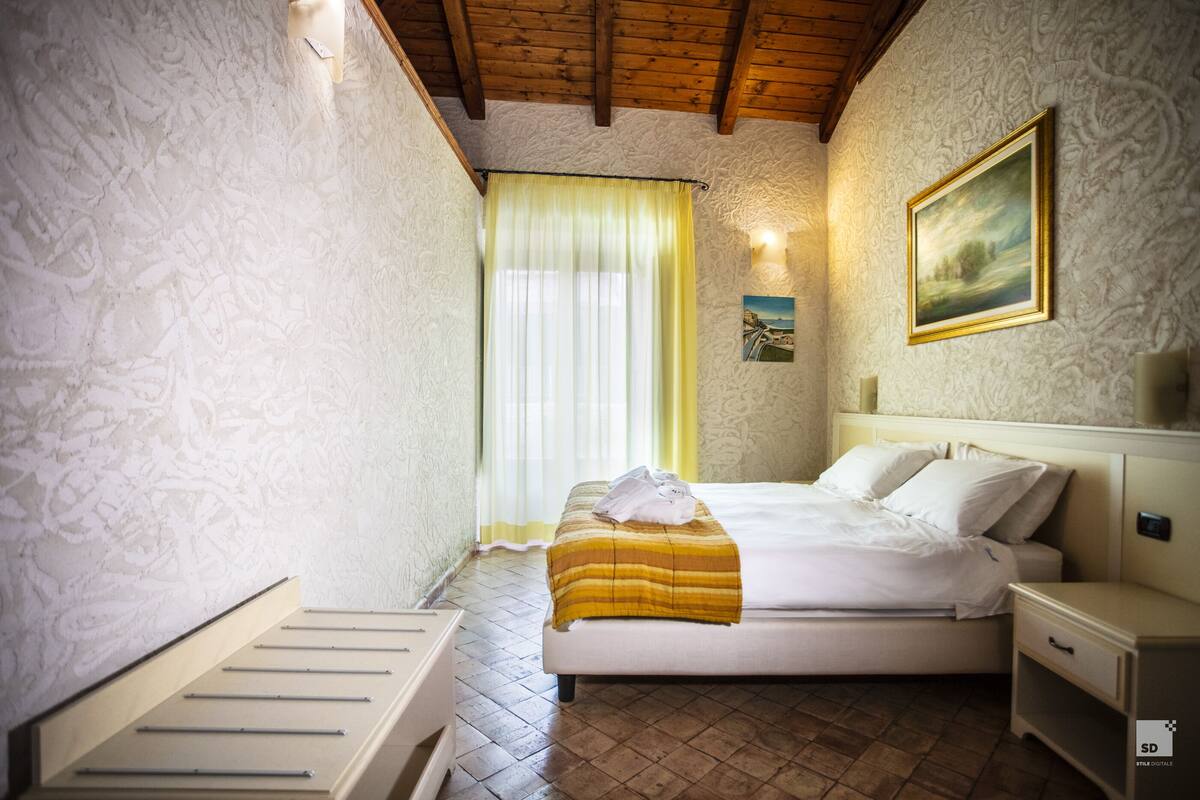 A bedroom with a bed, a nightstand, a bedside table, a window with curtains, and a painting on the wall.