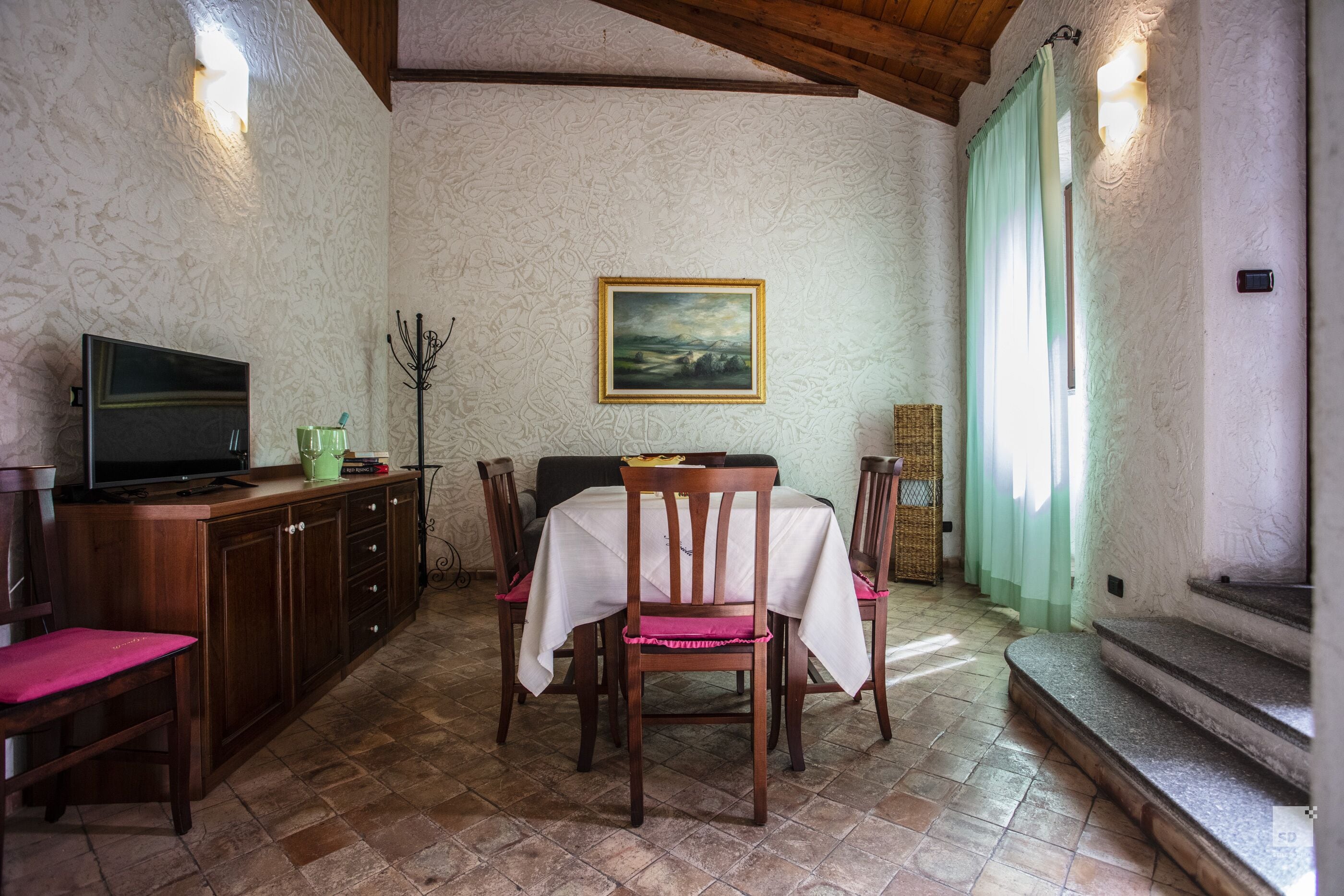 A dining area with a table set for two, a television on a cabinet, and a painting on the wall.
