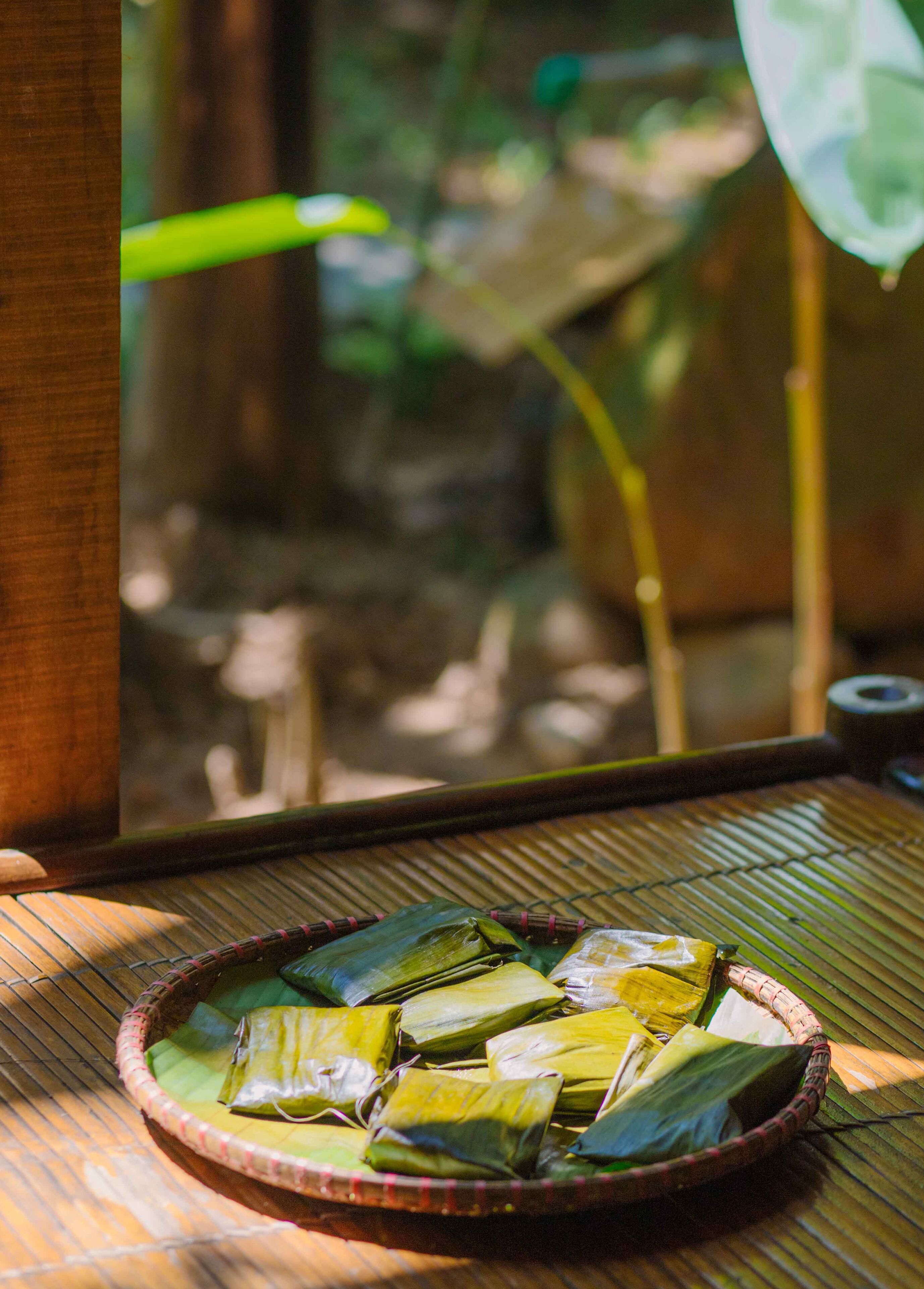 Un tapis en bambou sur lequel repose un panier tressé contenant plusieurs aliments emballés dans du papier vert et jaune, vraisemblablement des plats traditionnels, le tout posé sur une surface en bois.