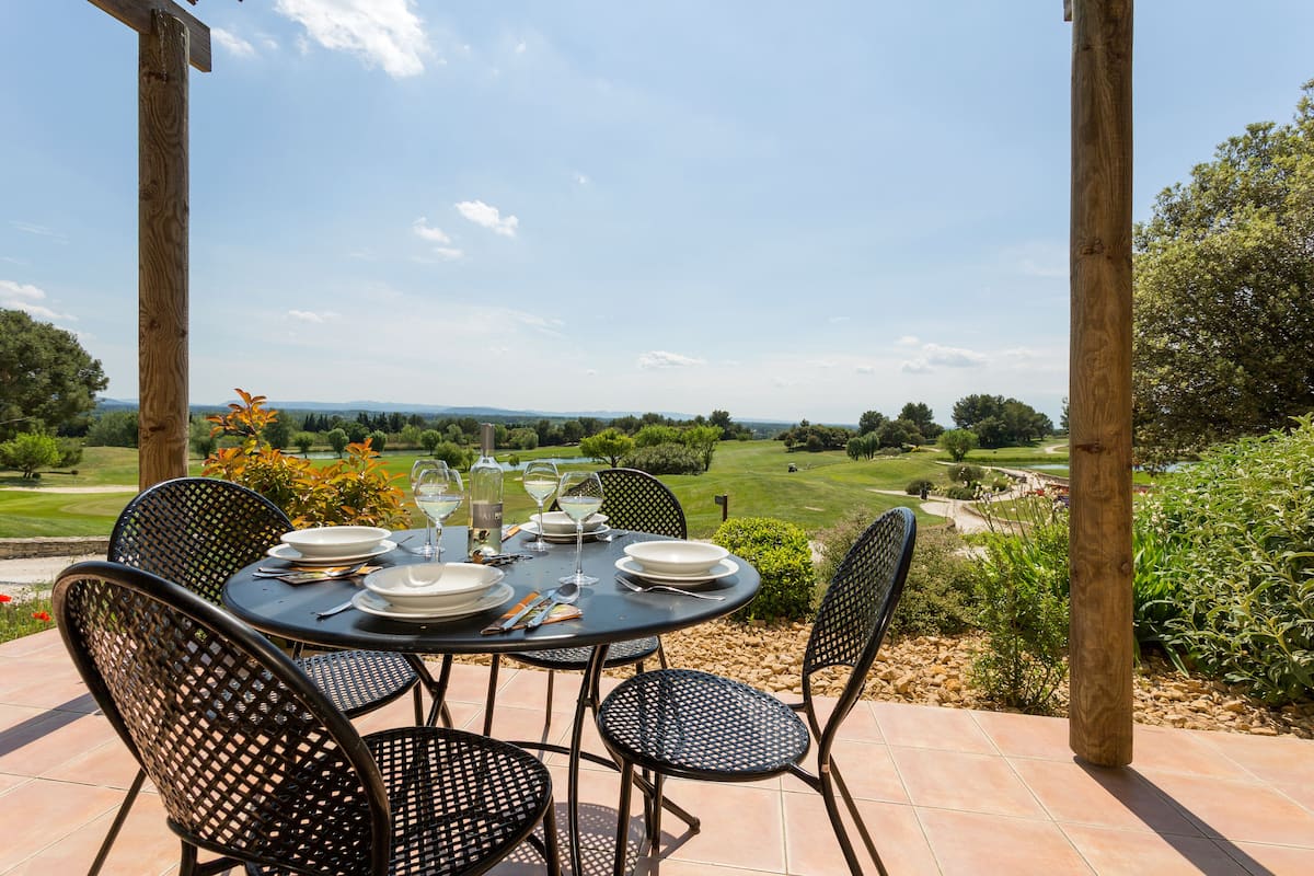 A round table set for four with plates, glasses, and cutlery, surrounded by black mesh chairs, overlooking a golf course.