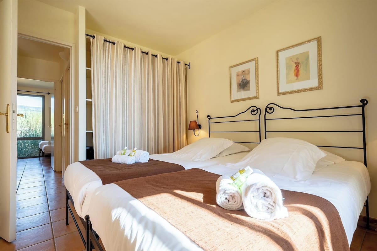 A hotel room with two beds, a view to the outside, and two framed pictures on the wall.