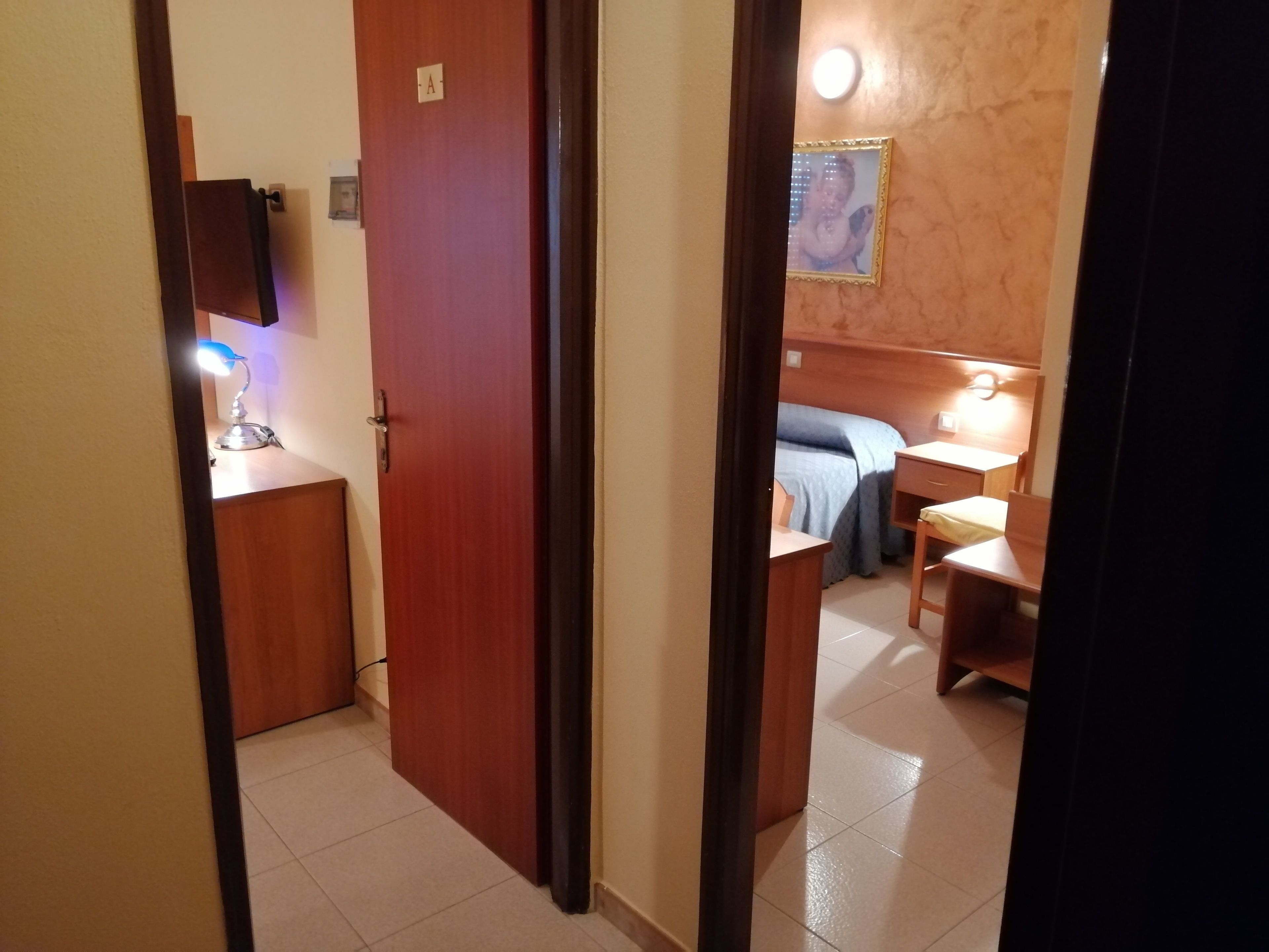 A hotel room with a wooden door, a desk with a lamp, a bed with a blue blanket, and a small table with a chair.