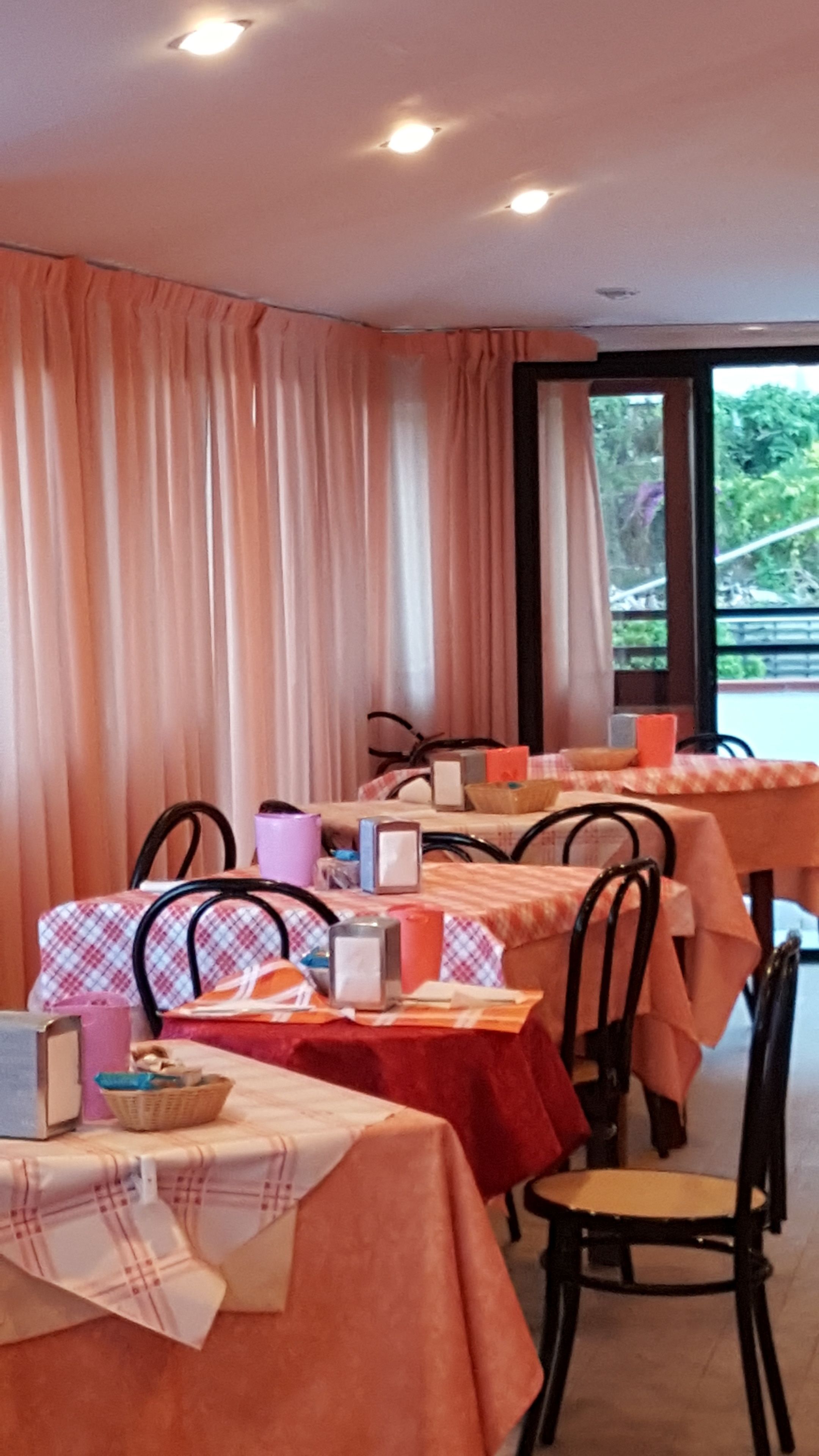 A restaurant interior with tables covered in checkered tablecloths, black metal chairs, and a large window with curtains.