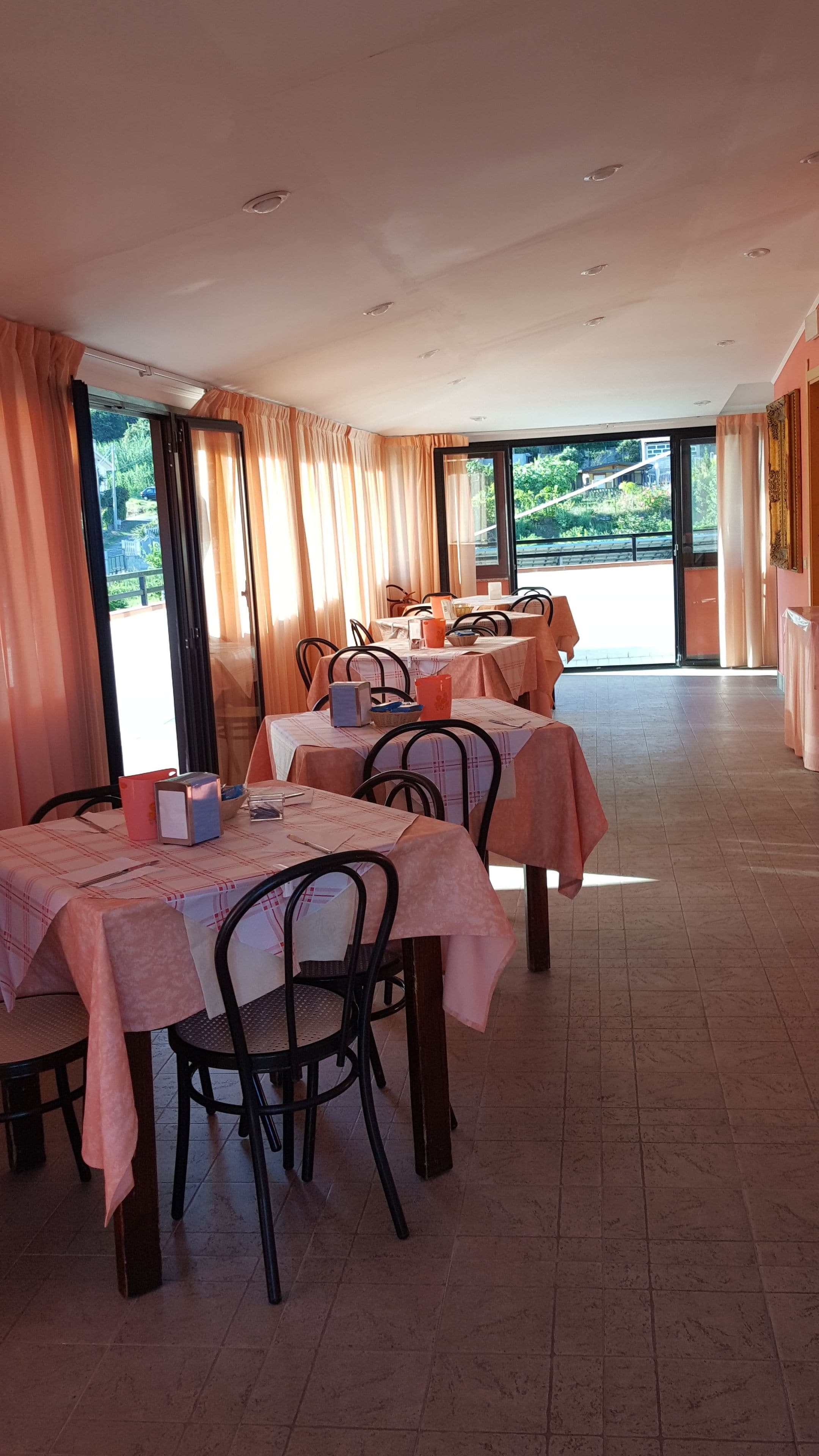 Un comedor con mesas dispuestas para el servicio, ventanas grandes con cortinas y vistas a zonas verdes al exterior.