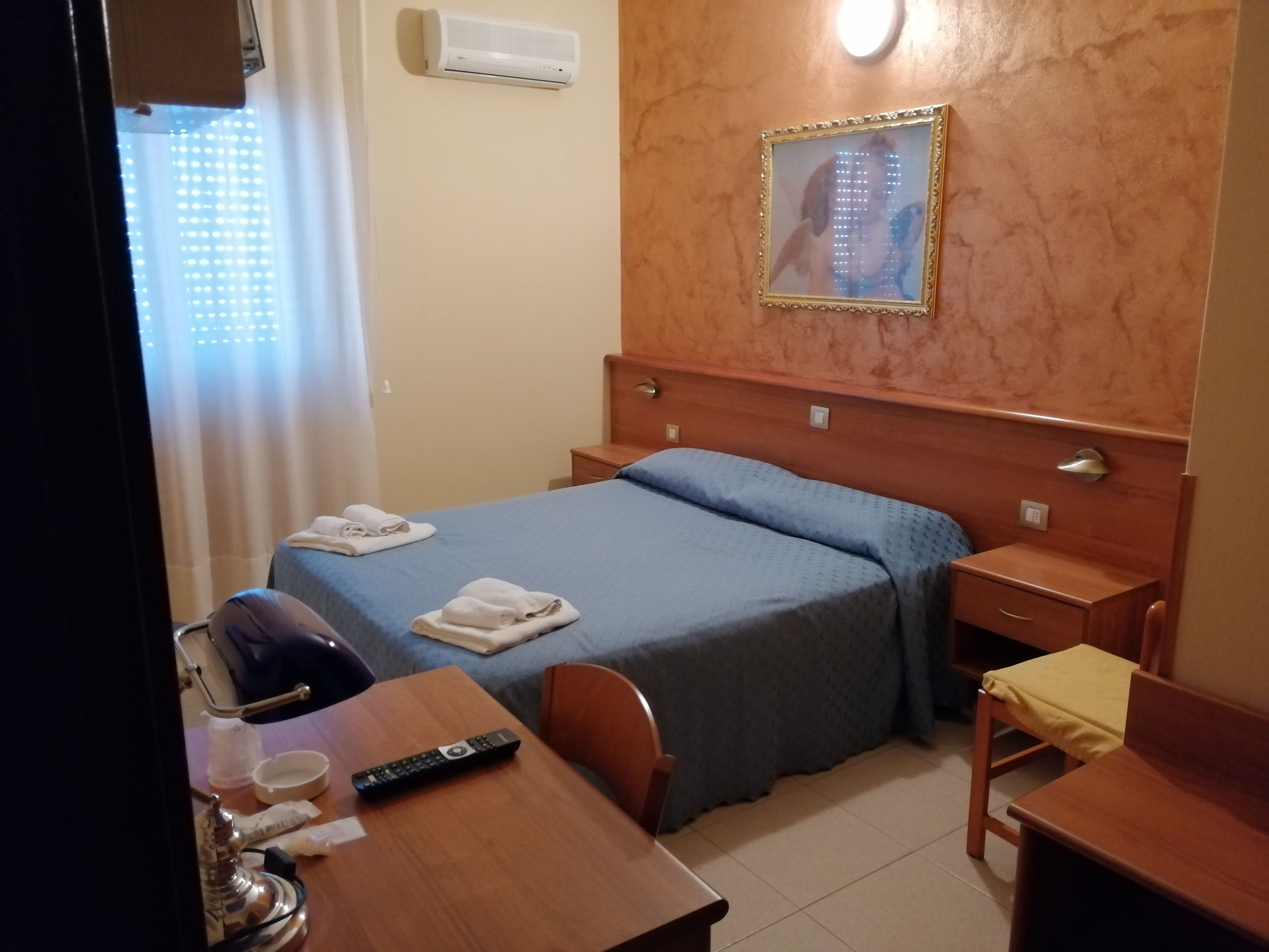 Habitación de hotel con cama, escritorio, silla, mesita de noche, aire acondicionado instalado en la pared y un cuadro enmarcado.