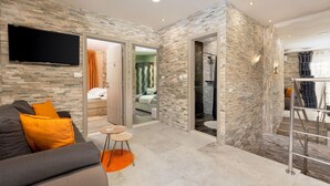 Un salon moderne avec un canapé gris, des coussins orange, un téléviseur à écran plat et un mur à la texture pierre.