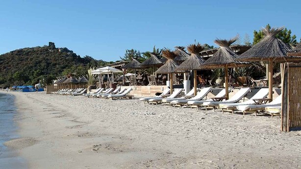 Een strand met ligbedden en rieten parasols, een heldere lucht en een heuvel met een gebouw op de top.