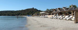 A beach with sun loungers and thatched umbrellas, a clear sky, and a hill with a structure on top. - Résidence Tamaris 2 (Lecci)
