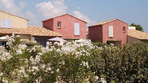 Een rij Mediterrane gebouwen met terracottadaken, witte luiken, en een tuin met bloeiende planten op de voorgrond.