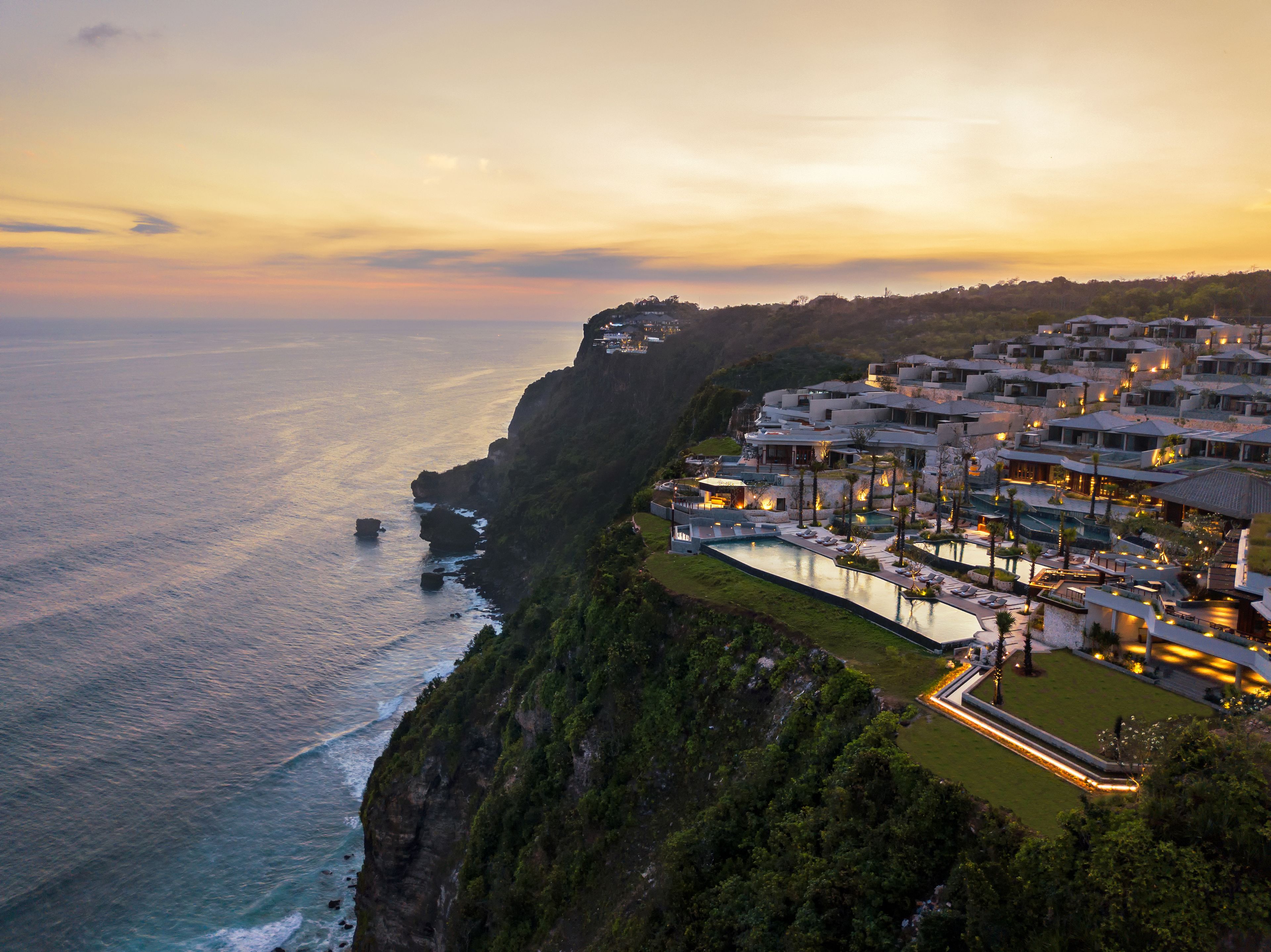 A coastal hotel with a cliffside view, multiple swimming pools, and a well-lit beachfront area.