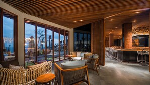 A modern interior with wicker furniture, a bar area, and large windows offering views of the cityscape. - Six Senses Bali (Pecatu)