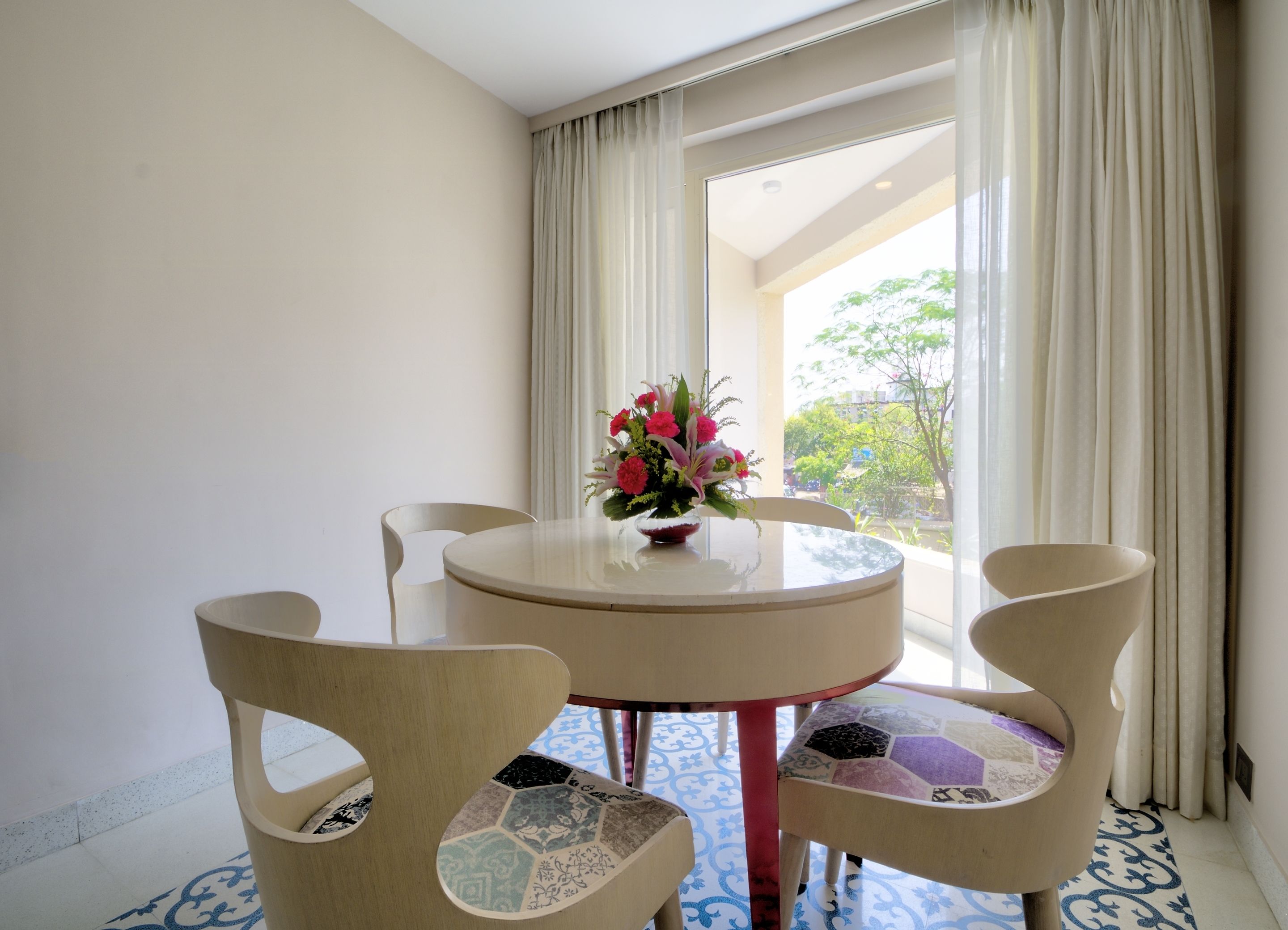 A round table with a vase of flowers, two chairs with patterned cushions, and a view of greenery through a window with curtains.