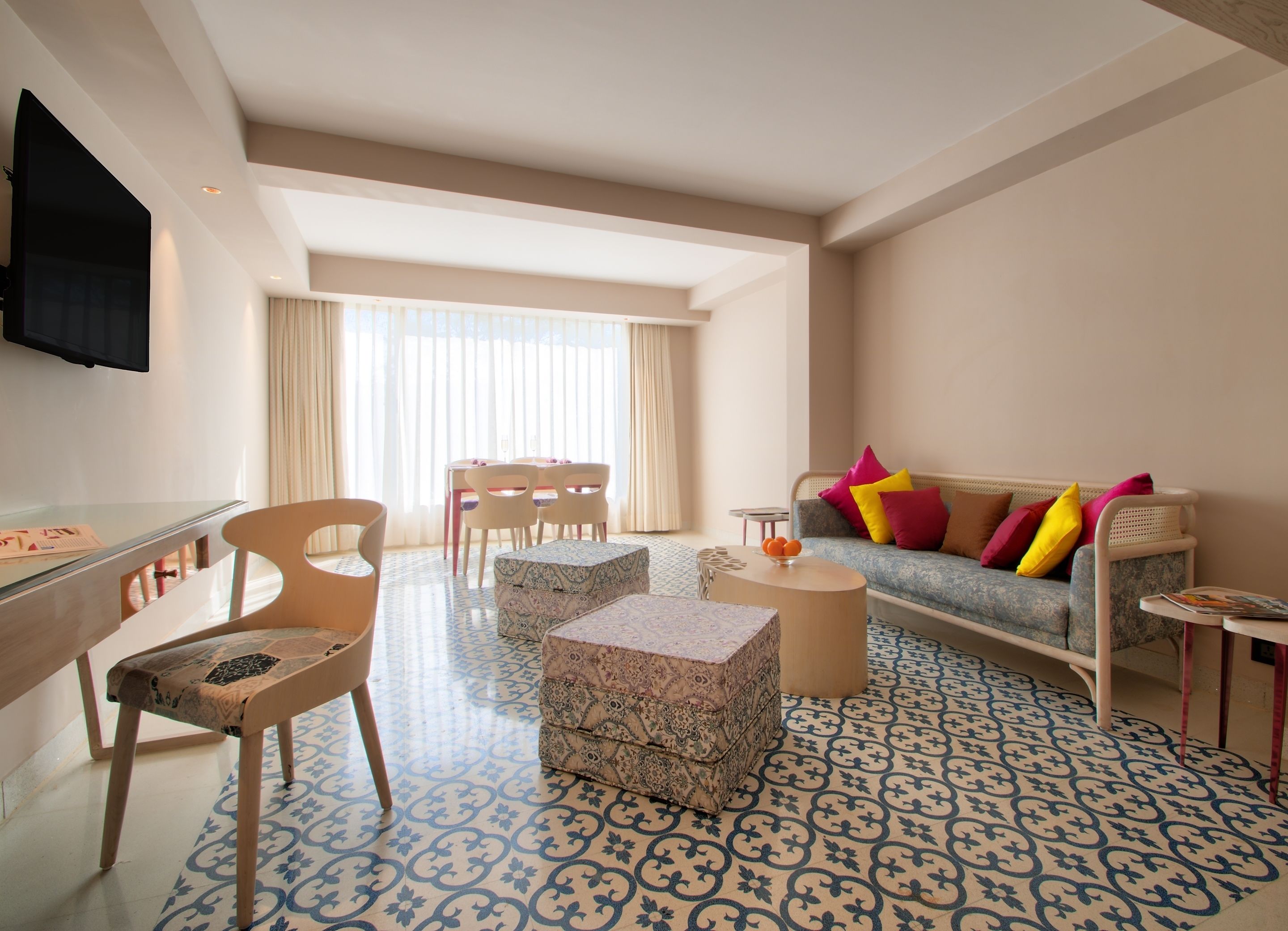 Une chambre d’hôtel moderne dotée d’un coin repas, d’un canapé agrémenté de coussins colorés, d’un téléviseur fixé au mur et de carreaux de sol à motifs.