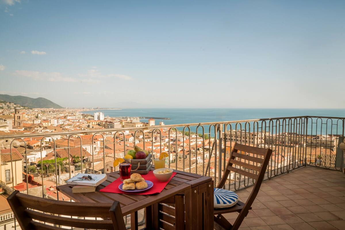 Varanda com mesa posta para o café da manhã, com vista para a paisagem urbana costeira.