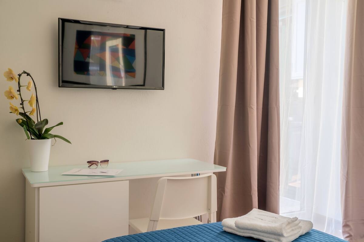 Quarto de hotel com cama, escrivaninha, cadeira, televisão na parede e janela com cortinas.