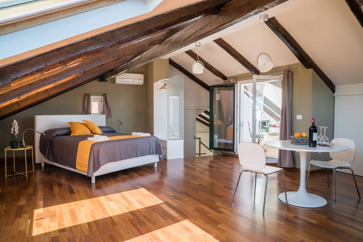 Quarto com teto inclinado, cama com colcha cinza e laranja, mesa redonda branca com cadeiras, e vista para o exterior através de uma porta de vidro.