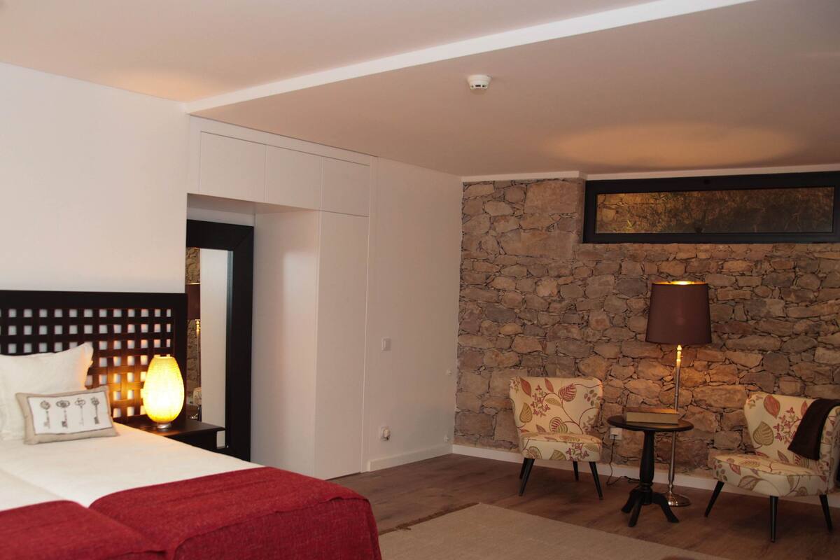 A hotel room with a bed, a nightstand, a lamp, two chairs, a small table, and a stone wall.