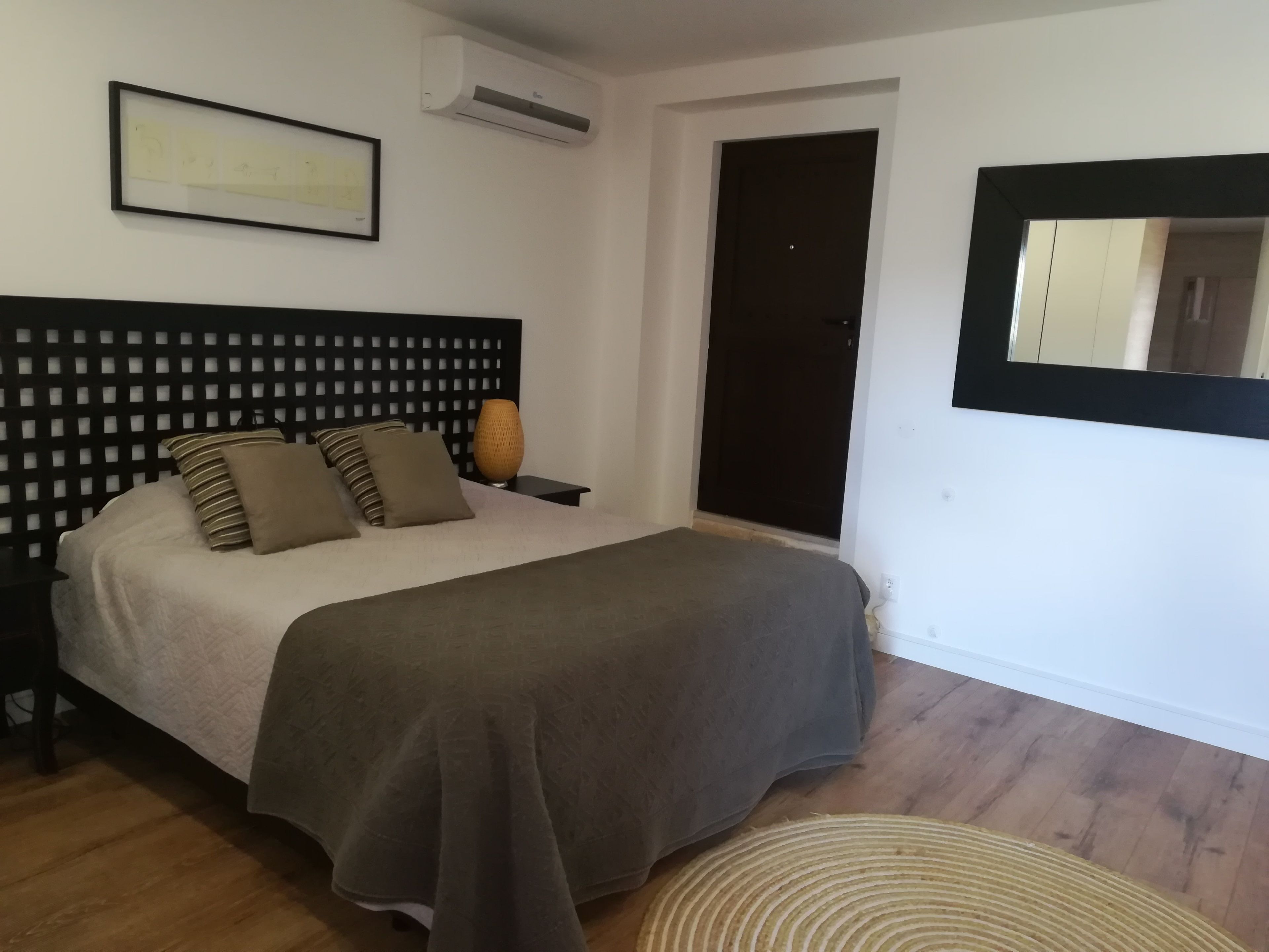 Une chambre à coucher avec un lit, une table de chevet, un climatiseur fixé au mur, un tableau encadré, une porte sombre et un miroir.