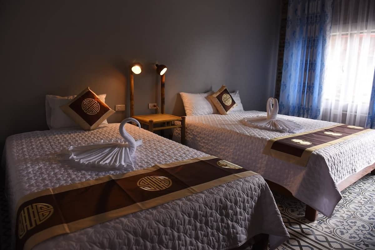Habitación de hotel con dos camas, cada una con un cojín decorativo con forma de cisne y una colcha estampada. Hay lámparas de pared y una mesita entre las camas.