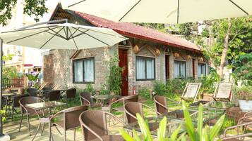 An outdoor patio with wicker chairs and tables, a brick building with a red-tiled roof, and a large umbrella providing shade.