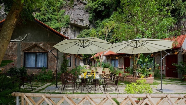 A rustic outdoor dining area with a brick building, two umbrellas, and a wooden fence. - Tam Coc Lakeside Homestay (Nam Hoa Lu)