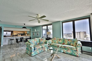 A spacious living room with a large window, a ceiling fan, and a kitchen area in the background.