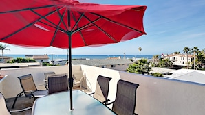 Une terrasse avec une table et des chaises, un parasol rouge, et une vue sur la mer et les bâtiments.