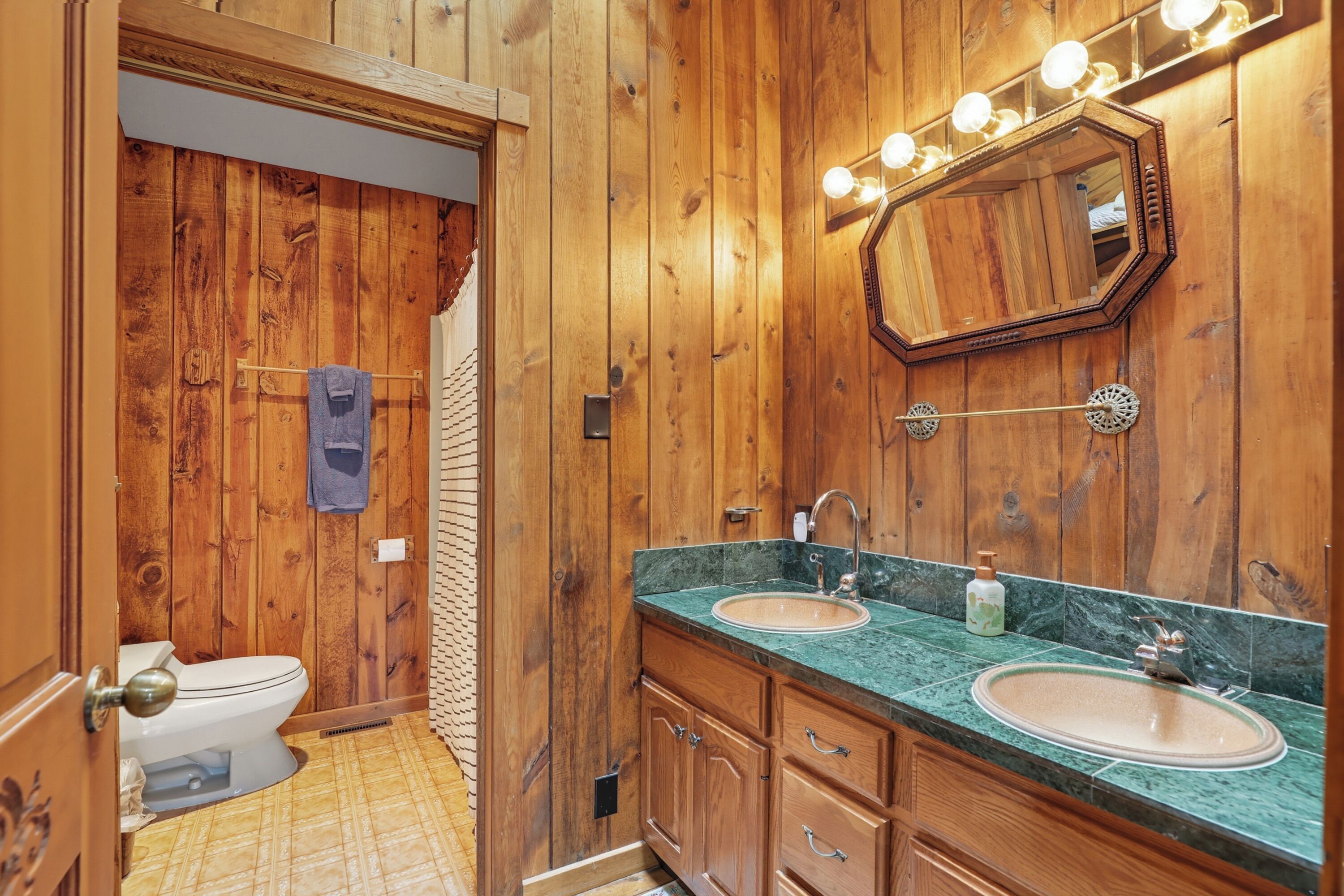 Un baño revestido de paneles de madera con dos lavamanos, un espejo con luces y un perchero para toallas.