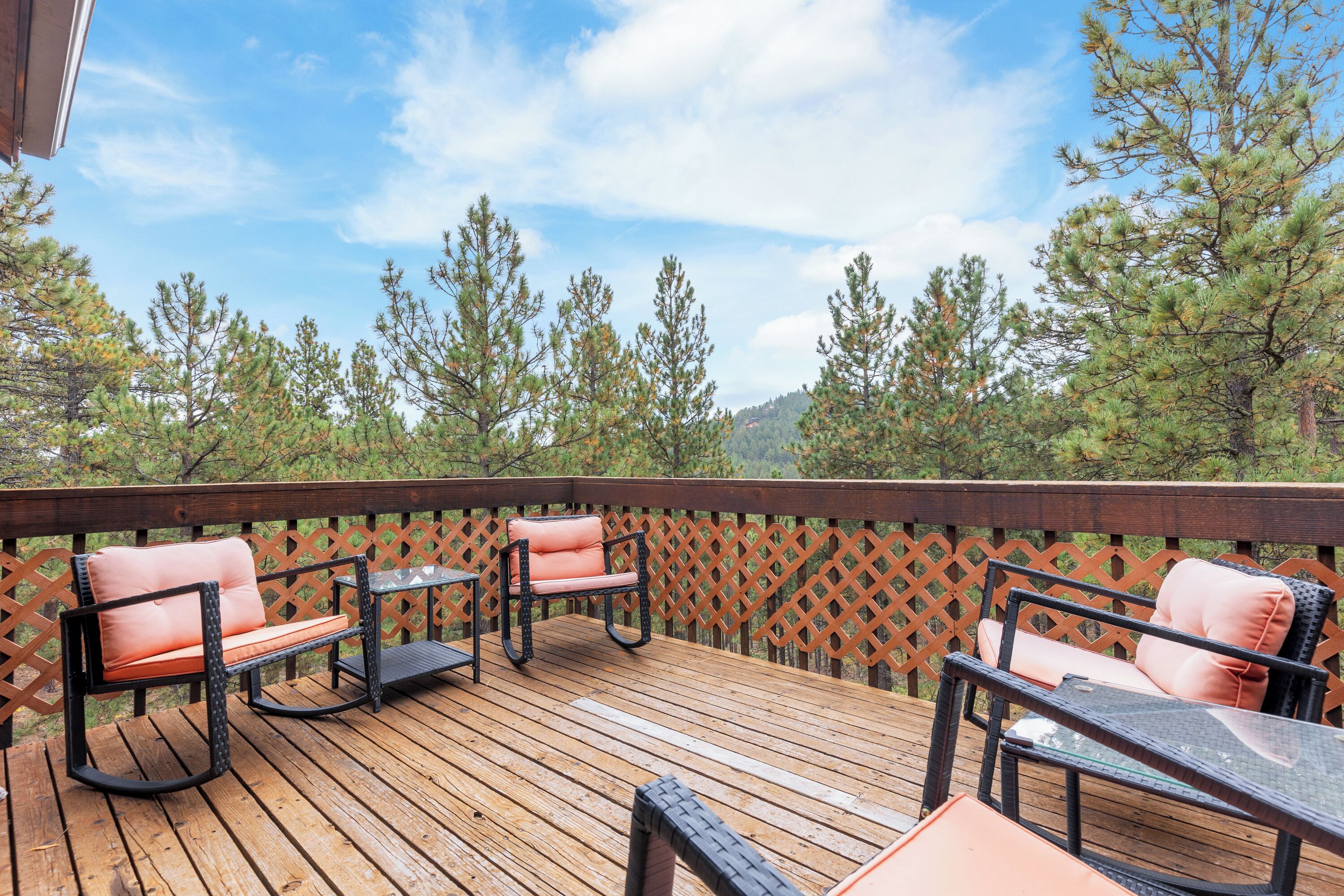 A wooden deck with outdoor furniture, including two cushioned chairs and a small table, surrounded by a lattice fence and overlooking a forested area.