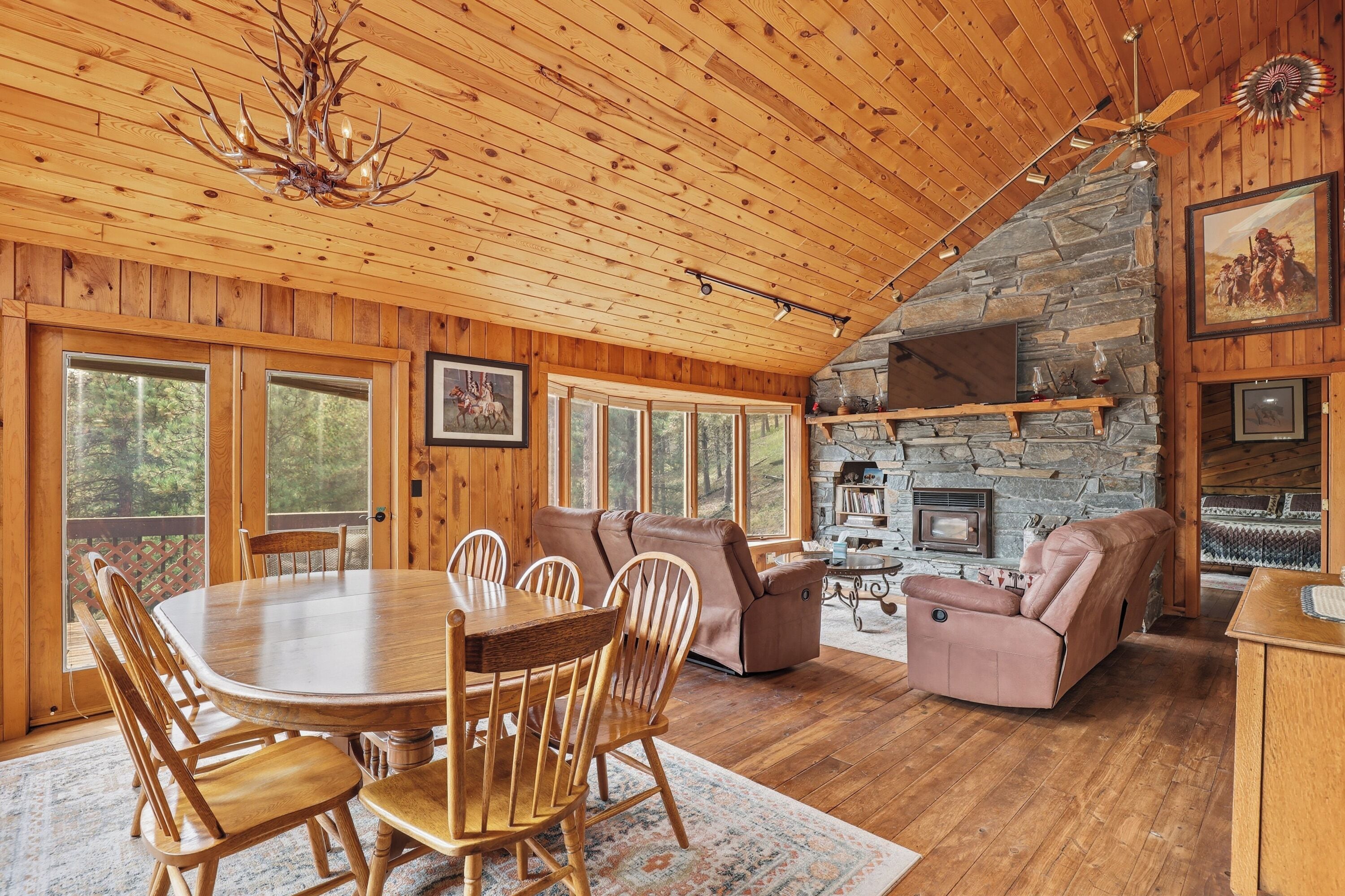A wooden cabin interior with a dining table, chairs, sofas, and a stone fireplace.