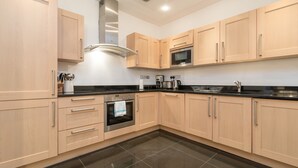 A modern kitchen with light wood cabinets, a stainless steel range hood, a built-in microwave, and a black countertop.