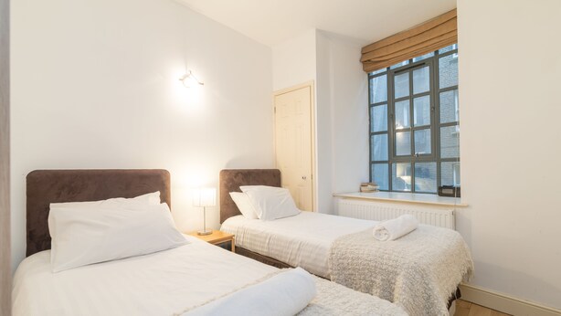 A hotel room with two beds, a window with curtains, a radiator, and a wall-mounted light fixture.