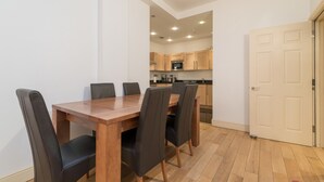 A dining area with a wooden table and black chairs, a kitchen in the background, and a white door.