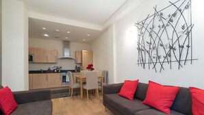 A modern living room with a grey sofa and red cushions, a dining area with a wooden table and chairs, and a kitchen in the background.