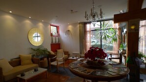 A well-lit living room with a round coffee table, sofas, and chairs. There is a large window with plants and a view of the outside. - Hotel Castillo del Alba (Somiedo)