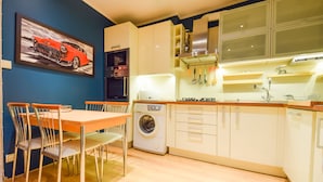 A compact kitchen with a washing machine, a dining table with chairs, and a painting of a car on the wall. - ShortStayPoland Leszno - B53 (Warsaw)