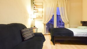 A room with a dark-colored sofa, a small table with a lamp, a bed with a green blanket, and a view of a blue-lit balcony through sheer curtains. - ShortStayPoland Leszno - B53 (Warsaw)