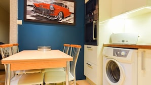 A compact kitchen with a washing machine, a dining table with chairs, and a framed picture on the wall. - ShortStayPoland Leszno - B53 (Warsaw)